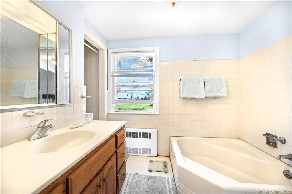 Bathroom with a white bathtub, wood vanity, cream tile, and a window with blinds.