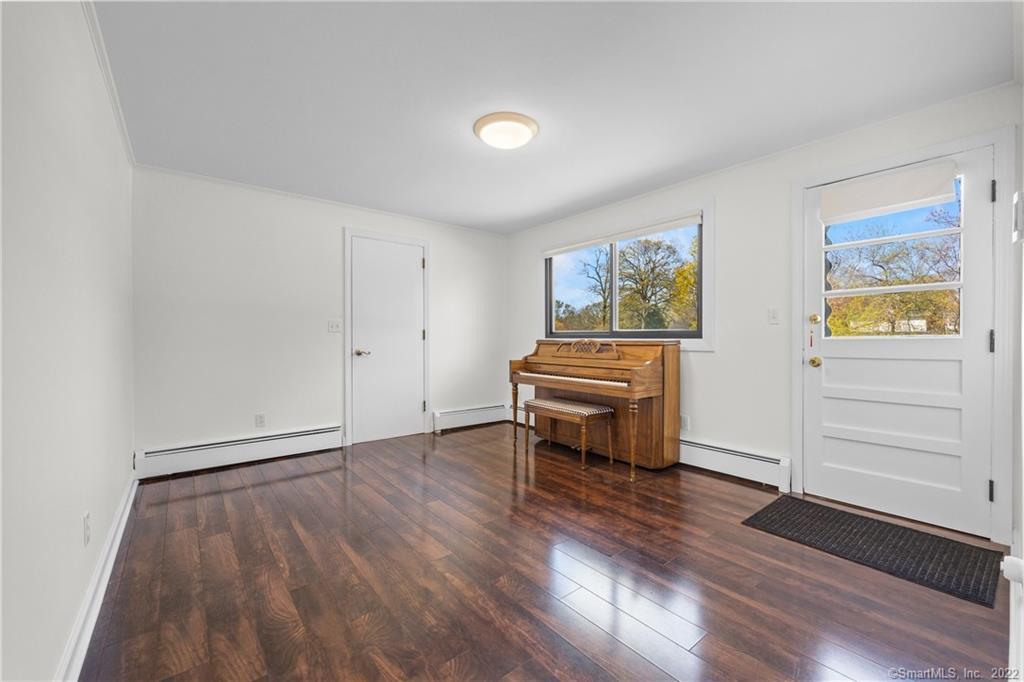 Room with an upright piano and bench, dark hardwood floors, and windows overlooking trees.