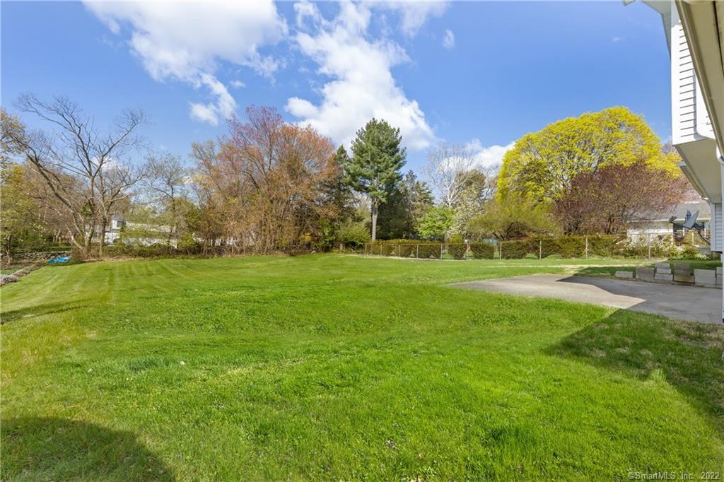 Backyard lawn bordered by mature trees and a fence, with a paved area beside the house.