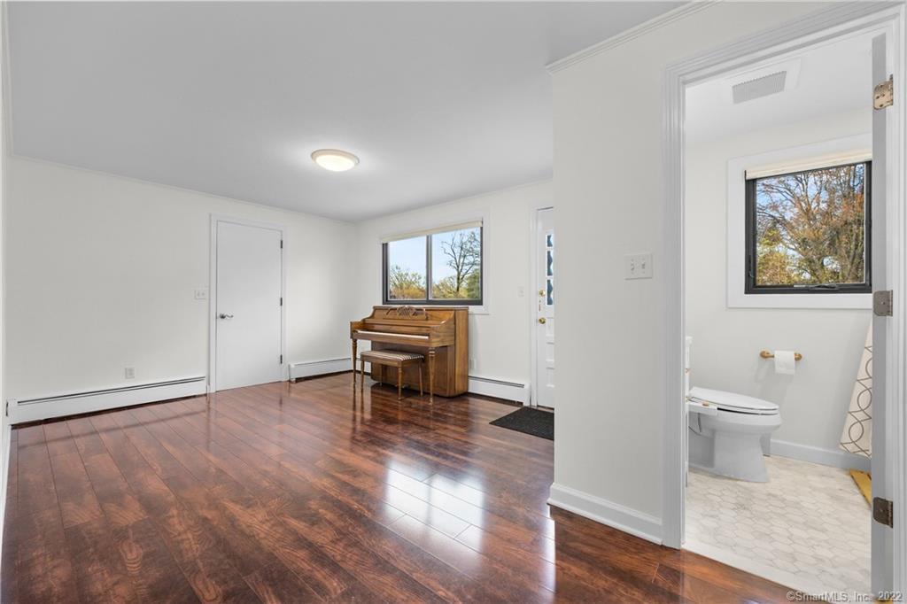 Bright room with an upright piano and hardwood floors, with an adjacent bathroom visible through the doorway.