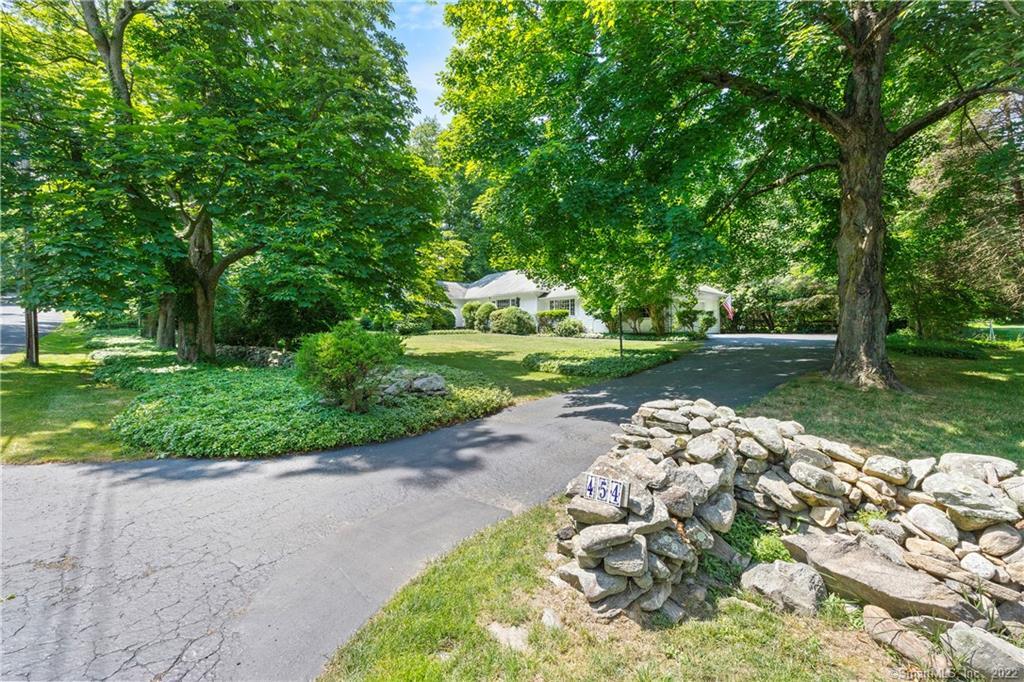 White home beneath mature trees, viewed from the curved driveway beside a stone wall marked 454.