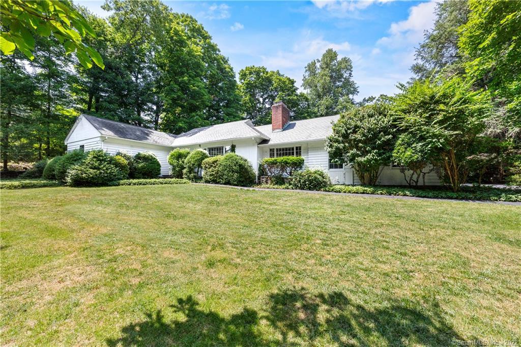 White ranch-style home with a brick chimney, broad lawn, and mature trees.