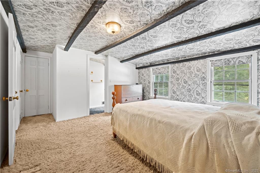 Bedroom with a large bed, patterned wallpaper and ceiling, dark beams, and two windows overlooking greenery.