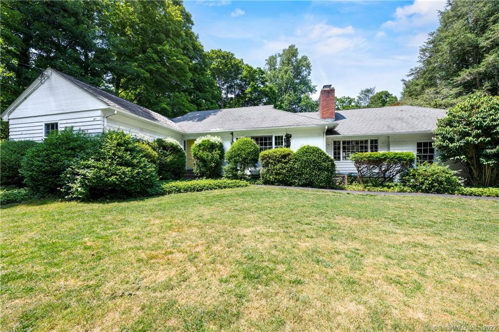 Single-story white house with a brick chimney, rounded shrubs, and a broad lawn.