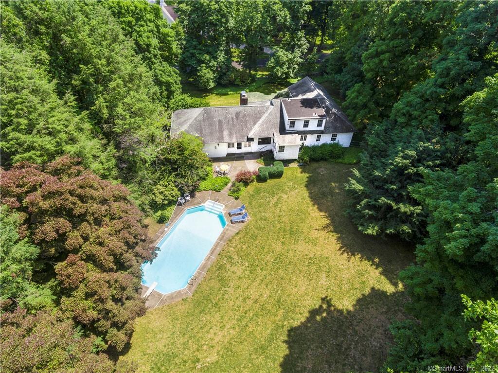 Aerial view of a white house, wooded lot, broad lawn, and in-ground swimming pool.