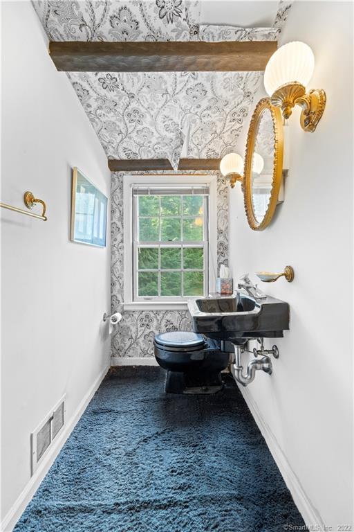 Powder room with a black toilet, blue carpet, patterned wallpaper, and an ornate brass mirror and sconce.