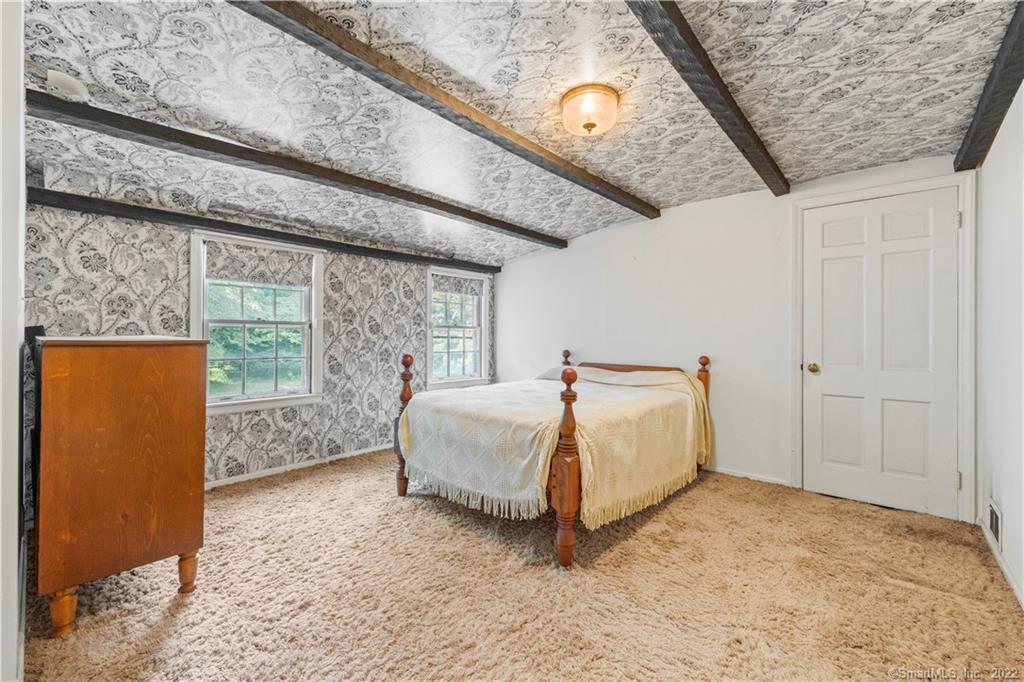 Bedroom with a four-poster bed, patterned ceiling and wallpaper, dark beams, and two windows.