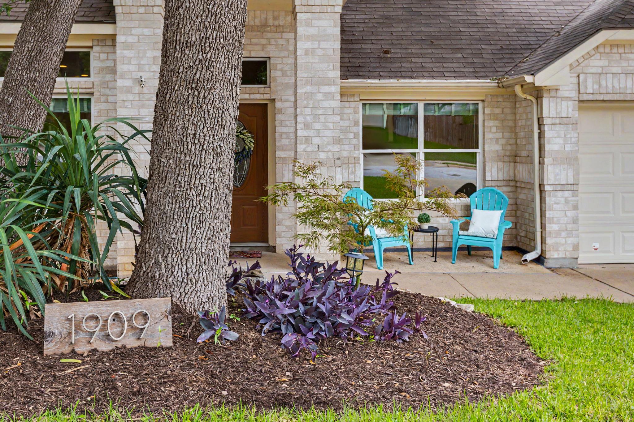 Stone house entrance framed by mature trees, with turquoise porch chairs and a 1909 address marker.