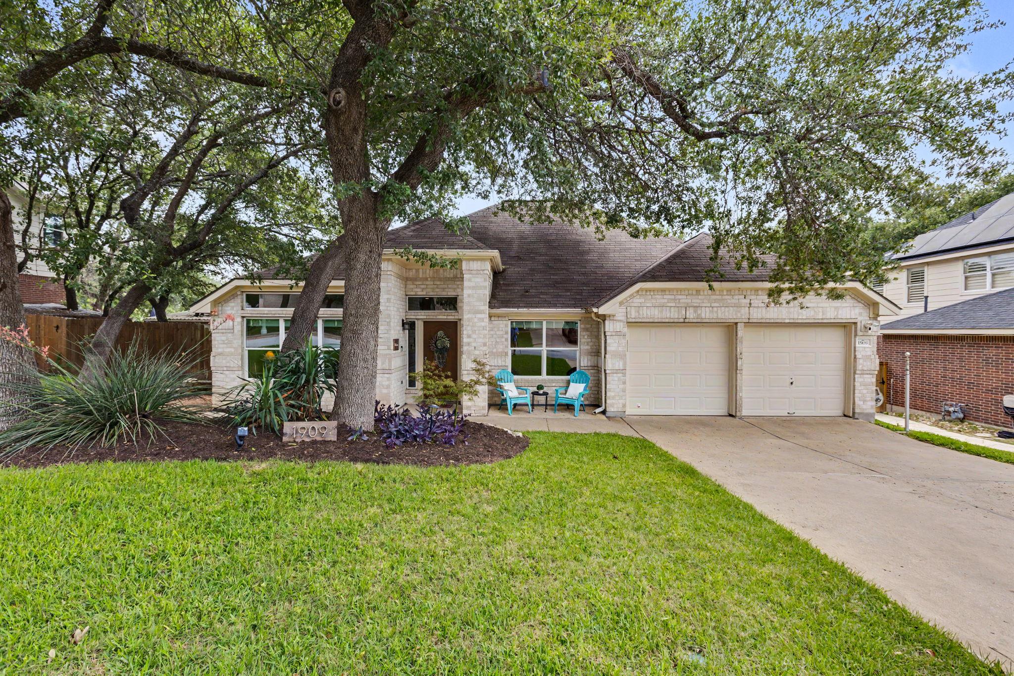 Stone single-story home with a two-car garage, mature shade trees, and turquoise chairs by the entrance.