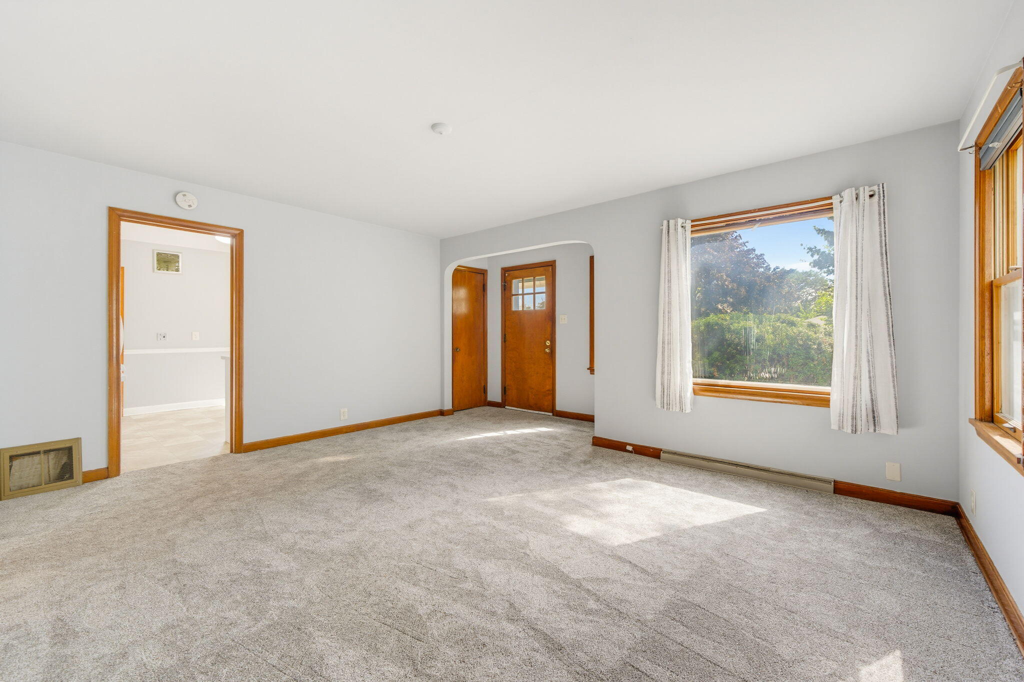 Empty living room with beige carpet, wood trim, and sunlight streaming through curtained windows.