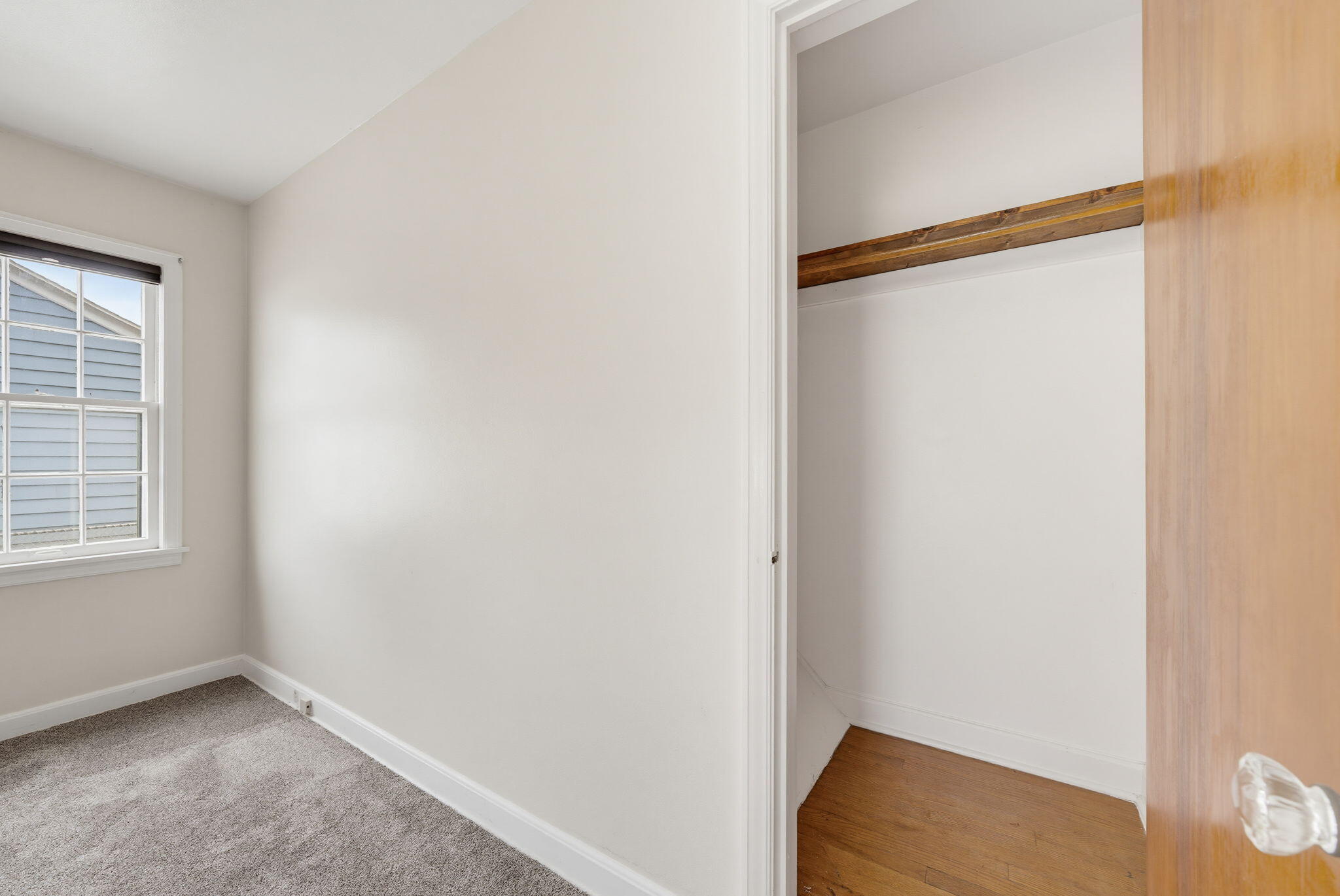 Empty bedroom with gray carpet and an open closet with hardwood flooring and a wooden shelf.