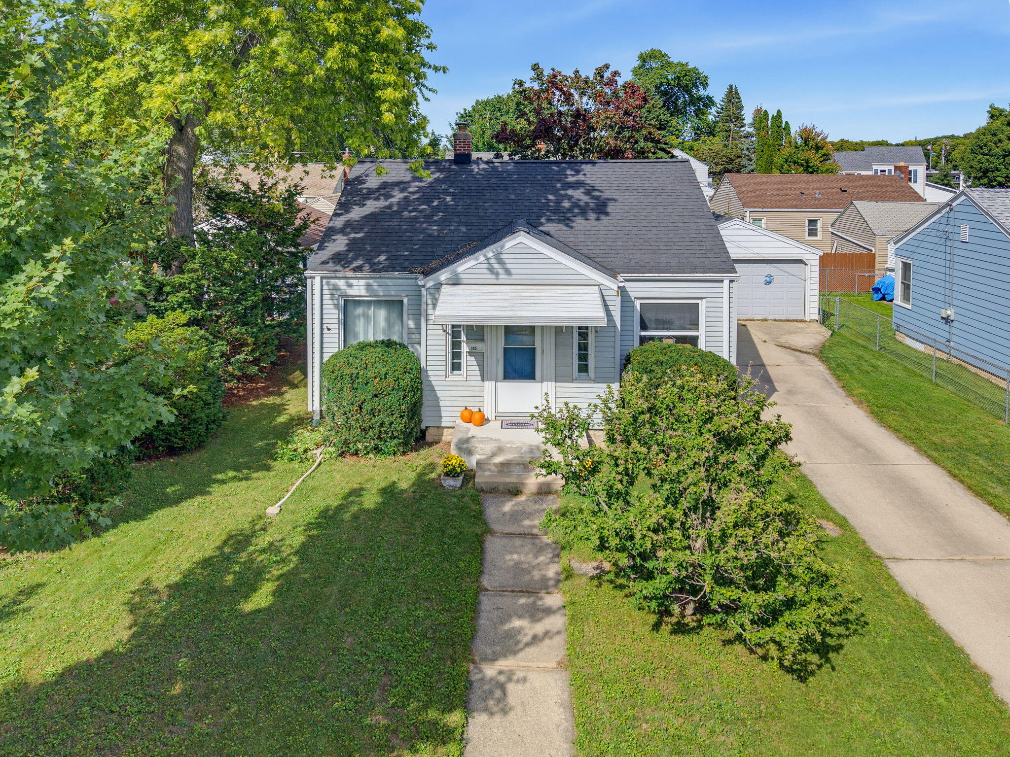 Light gray house with a blue front door, leafy shrubs, pumpkins, and a driveway.