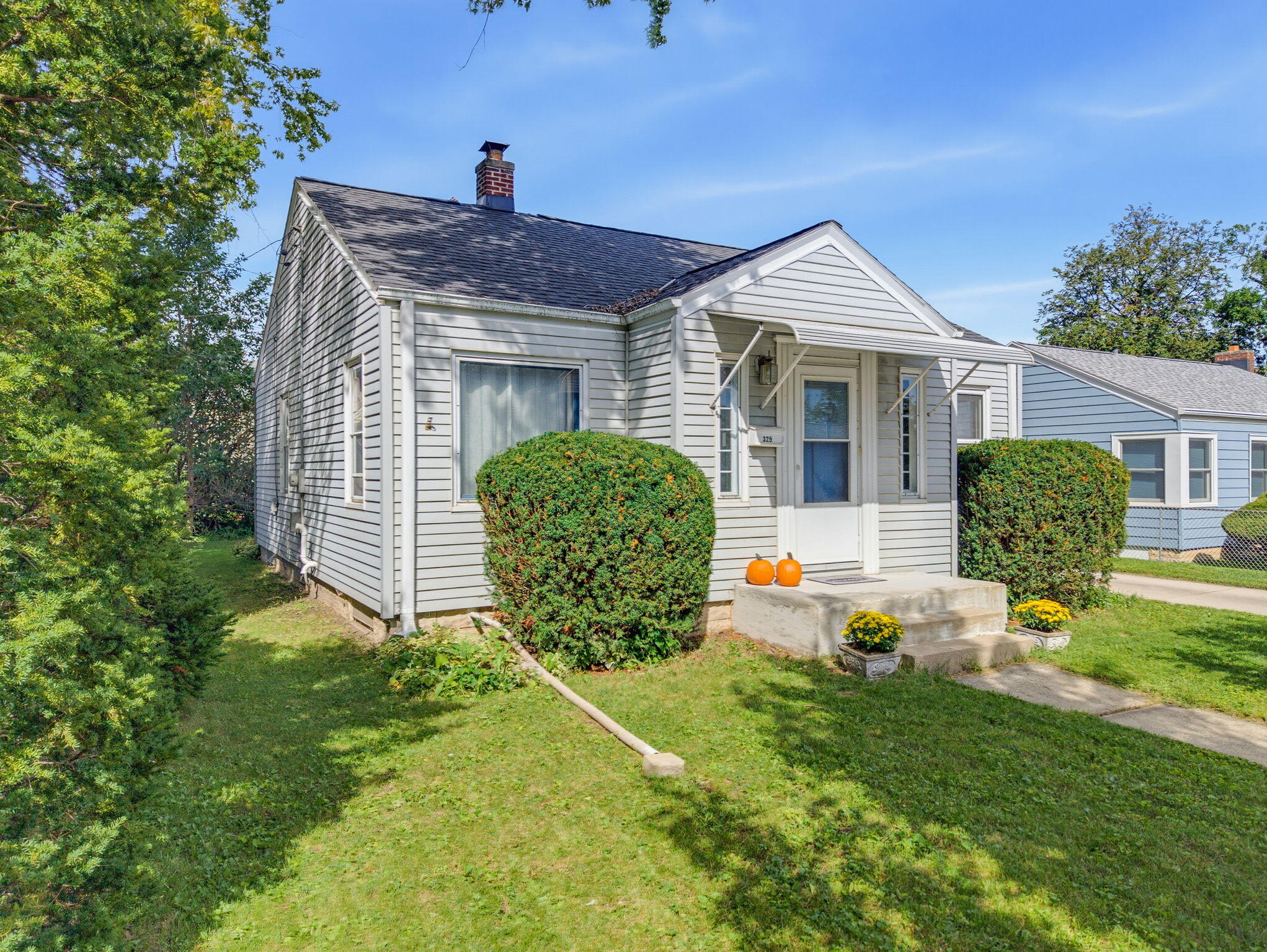 Light-colored clapboard house with a covered entrance, trimmed hedges, and two pumpkins on the front steps.