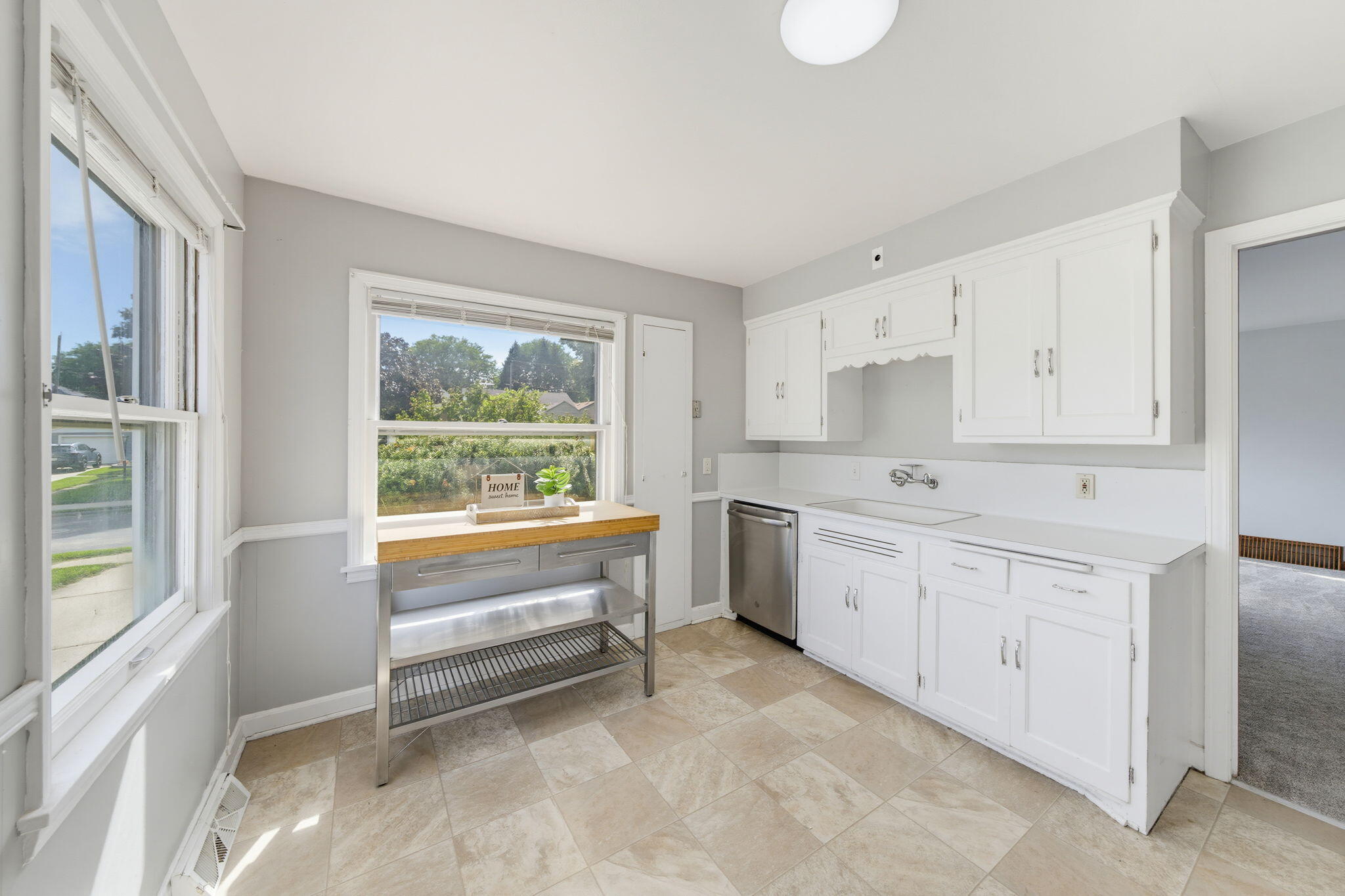 Bright kitchen with white cabinets, beige tile floors, and a metal worktable beneath a large window.