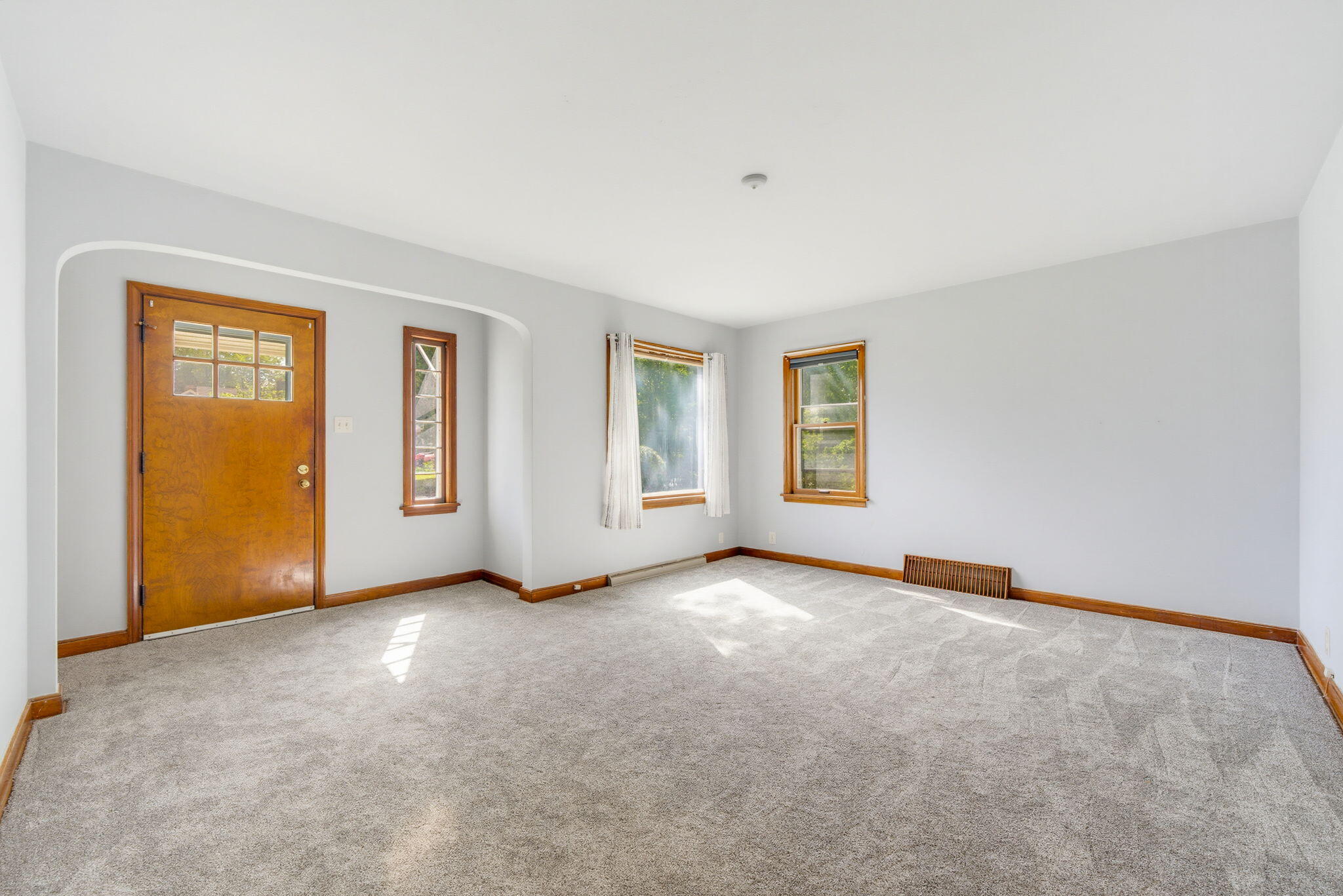 Empty carpeted room with a wood entry door, three windows, and sunlight across the floor.