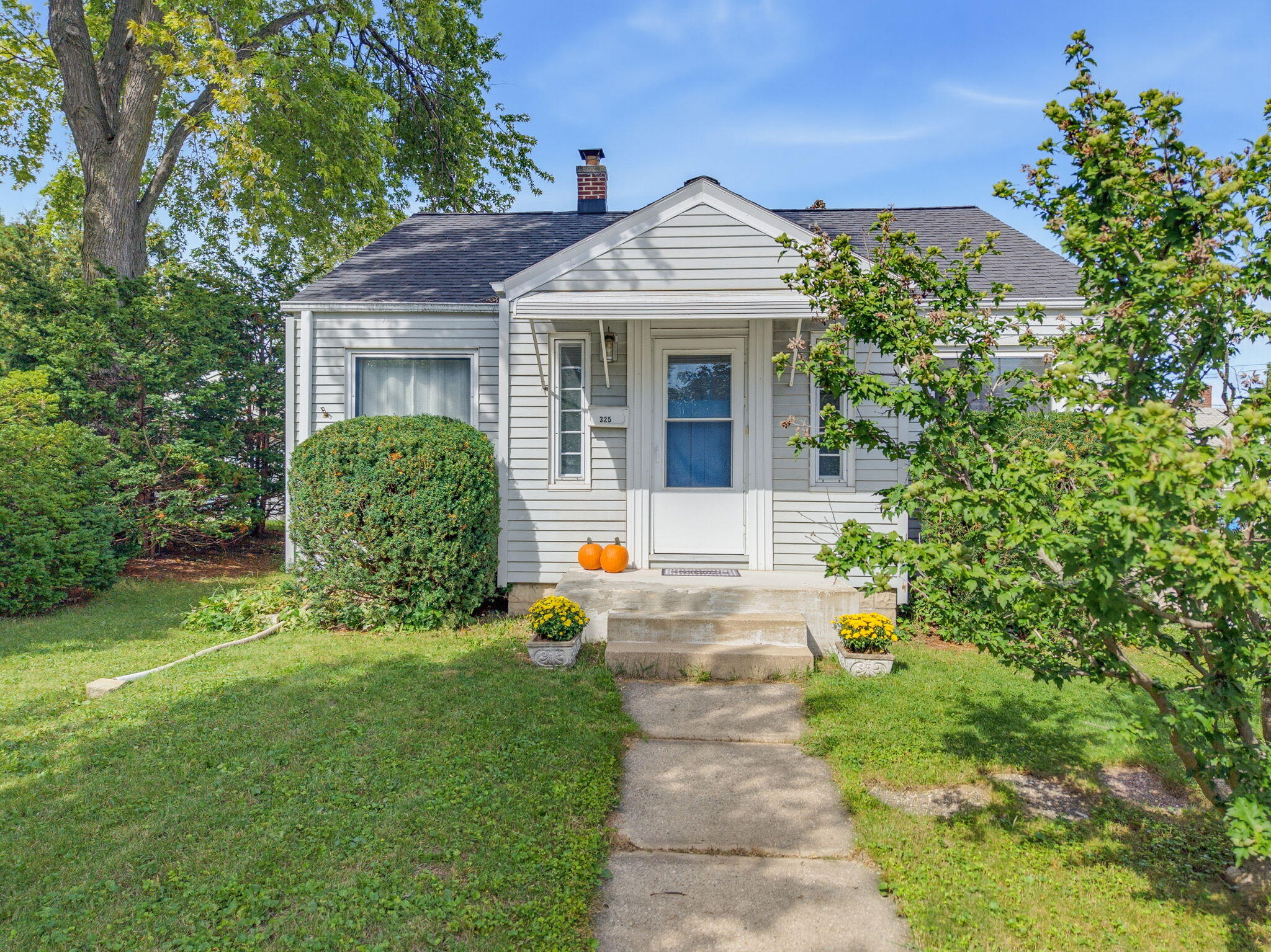 Light-gray house with a covered entry, front walkway, landscaped lawn, and pumpkins by the door.