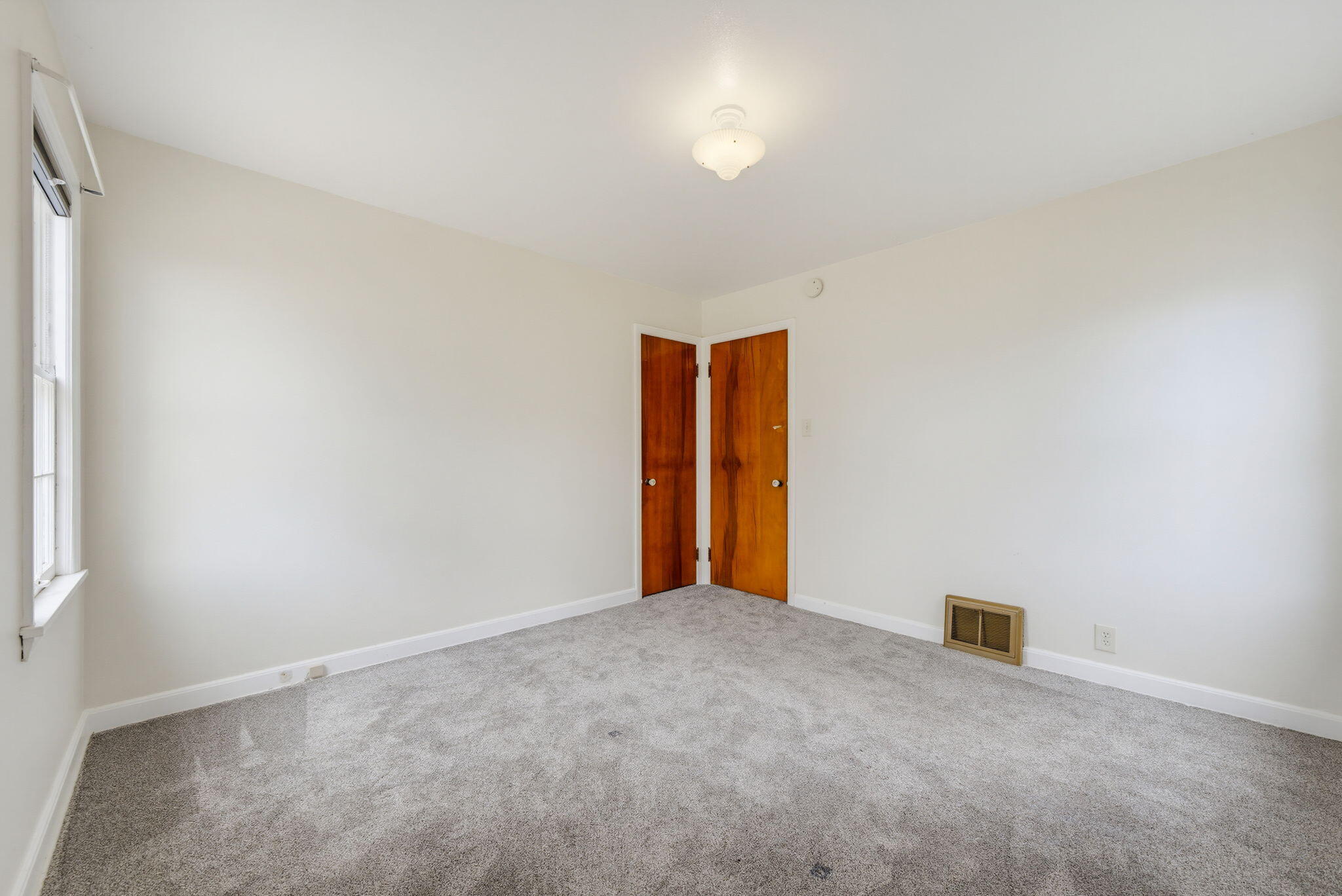 Empty carpeted room with a window and two wooden doors.