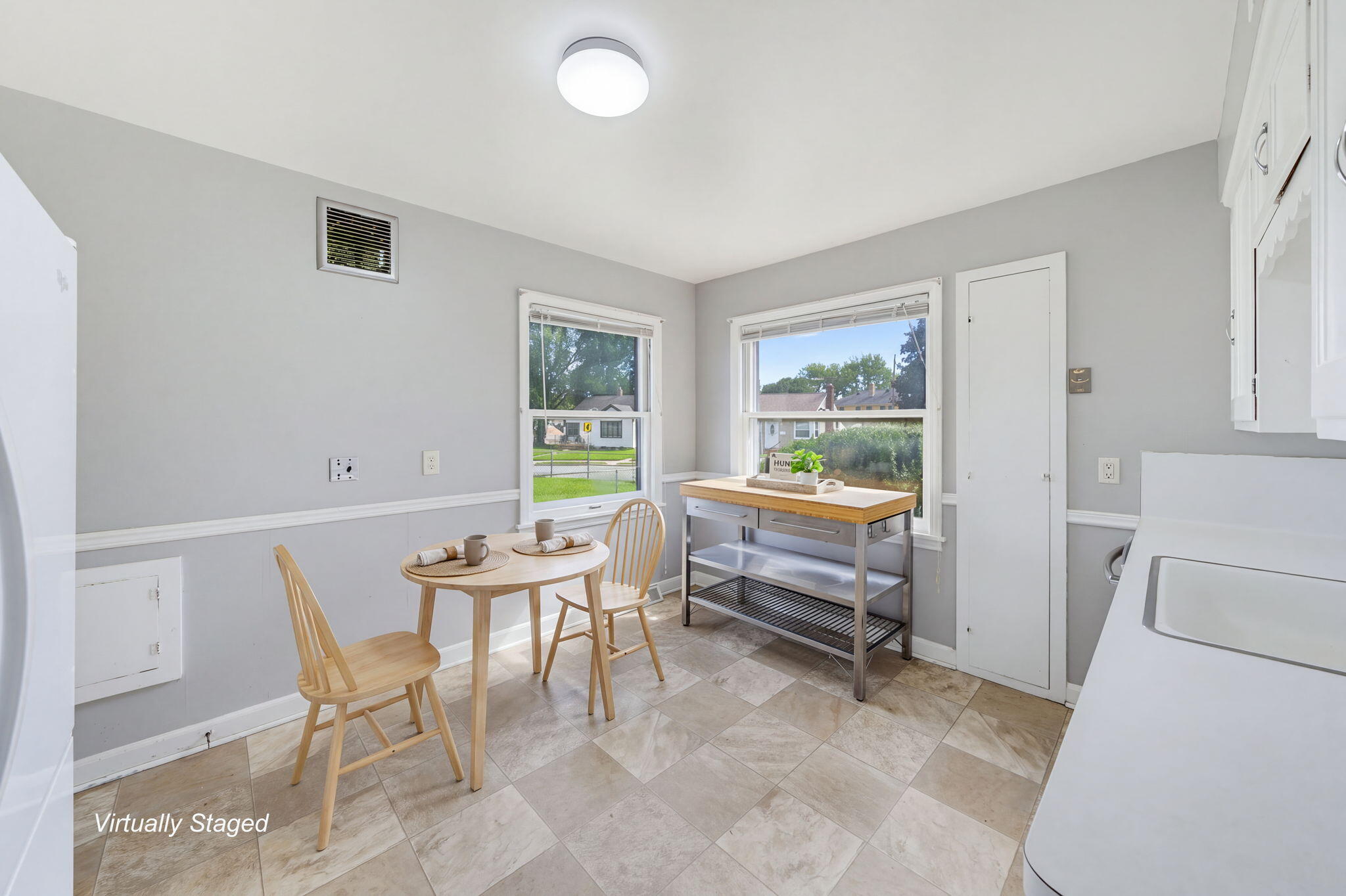 Virtually staged kitchen with a round wooden dining table, stainless prep table, and windows overlooking the street.