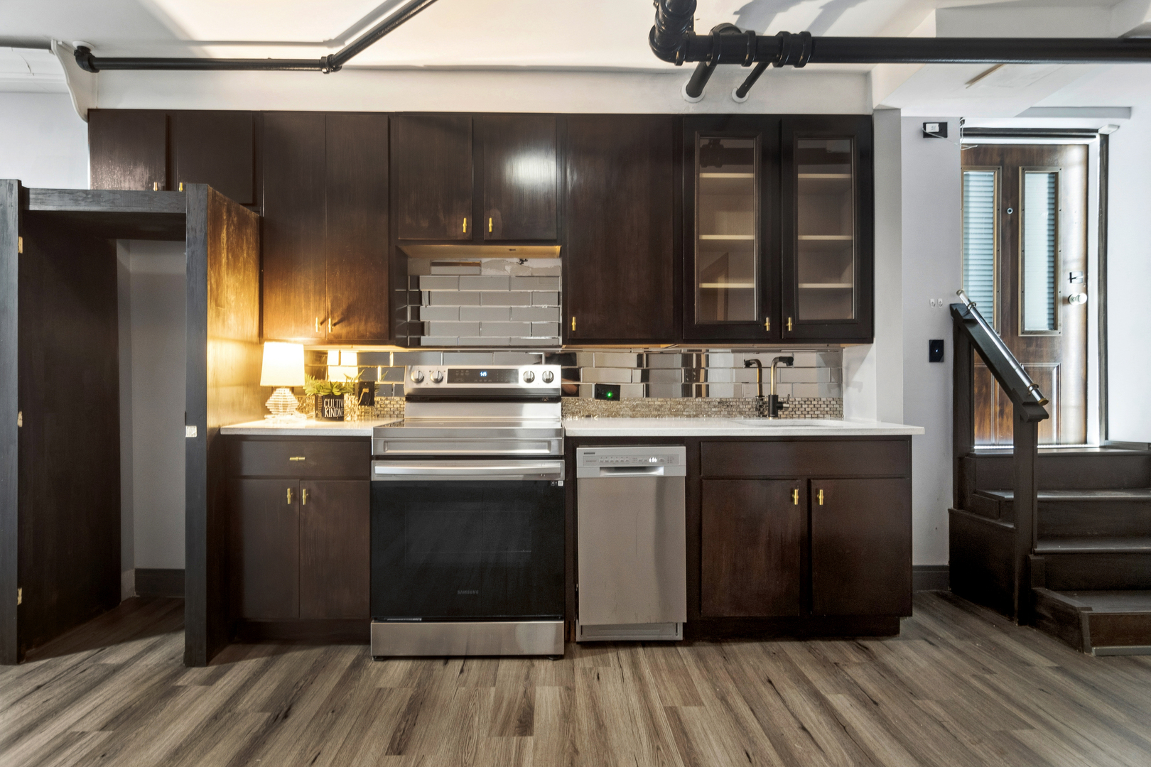 Kitchen with dark wood cabinets, stainless steel appliances, and a gray tile backsplash beside a staircase.