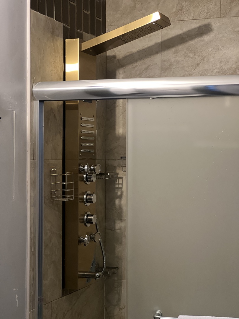 Glass-enclosed shower with a rainfall showerhead and a tall panel of body jets against beige stone tile.