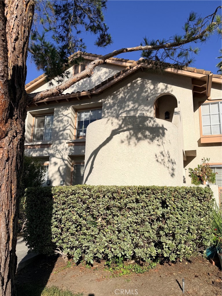 Two-story stucco home with a rounded balcony and tiled roof, partly shaded by a large pine tree.