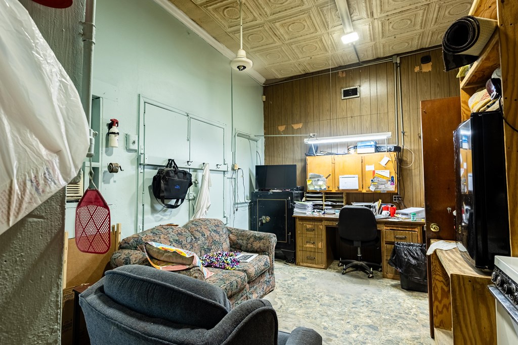 Cluttered office with a desk, computer, upholstered seating, wood-paneled walls, and a pressed-tin ceiling.