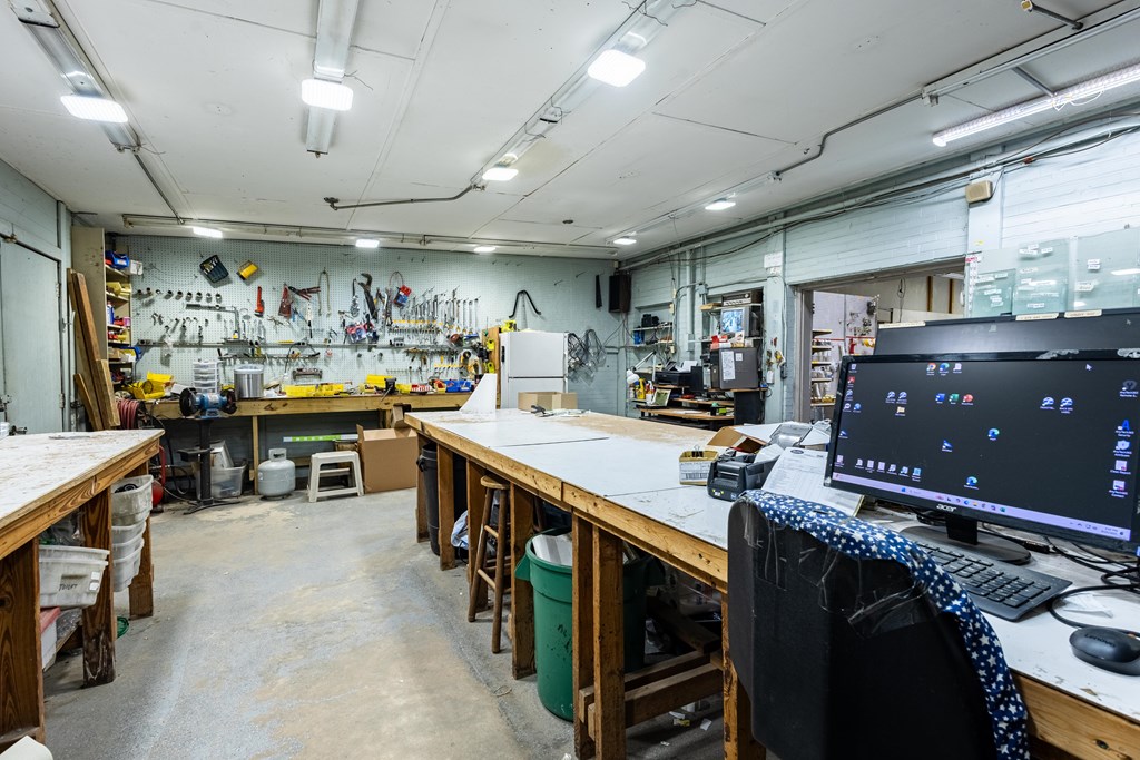 Workshop with tool-lined walls, long workbenches, storage bins, and a desktop computer.