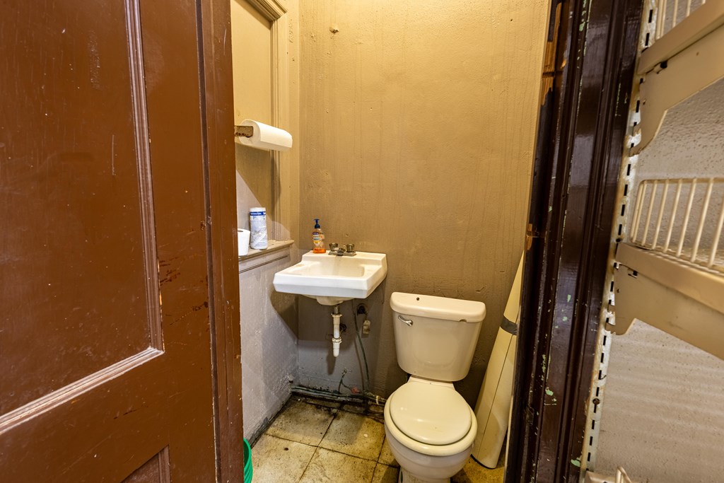 Small bathroom with a toilet, wall-mounted sink, exposed plumbing, and worn tile floor.