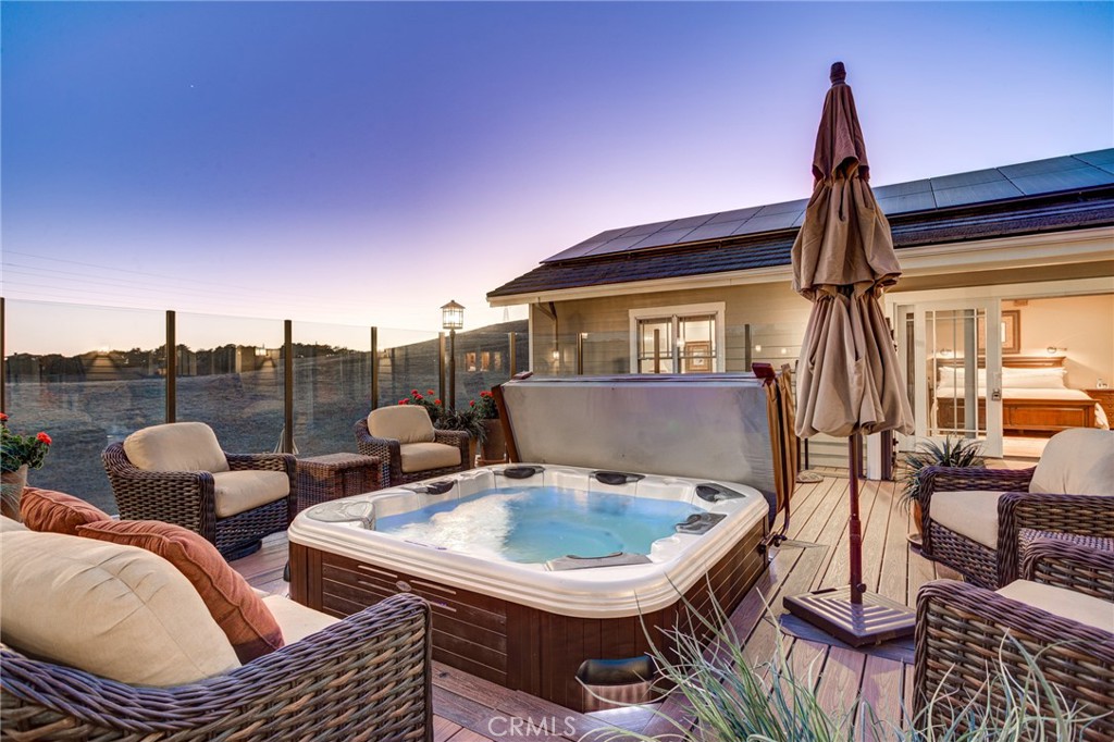Outdoor deck with an illuminated hot tub, wicker seating, and glass railing at dusk.