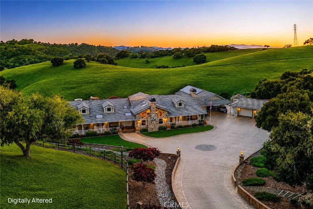 Aerial view of a stone-accented home and curved driveway amid rolling green hills at sunset.