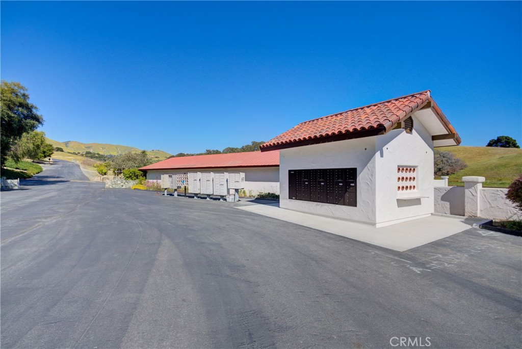 White community mailbox building with a red tile roof beside a paved road and rolling hills.