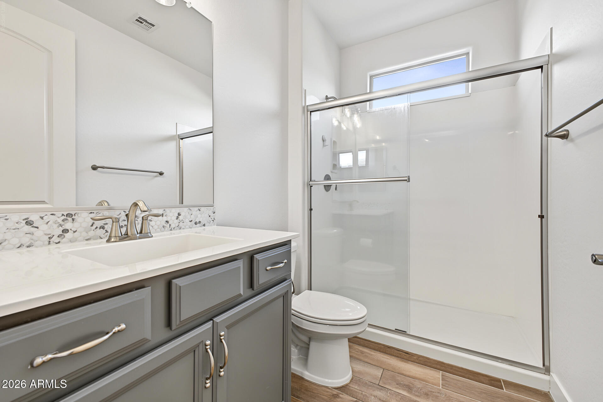 Bathroom with gray vanity, pebble-mosaic backsplash, and glass-enclosed shower beneath a high window.