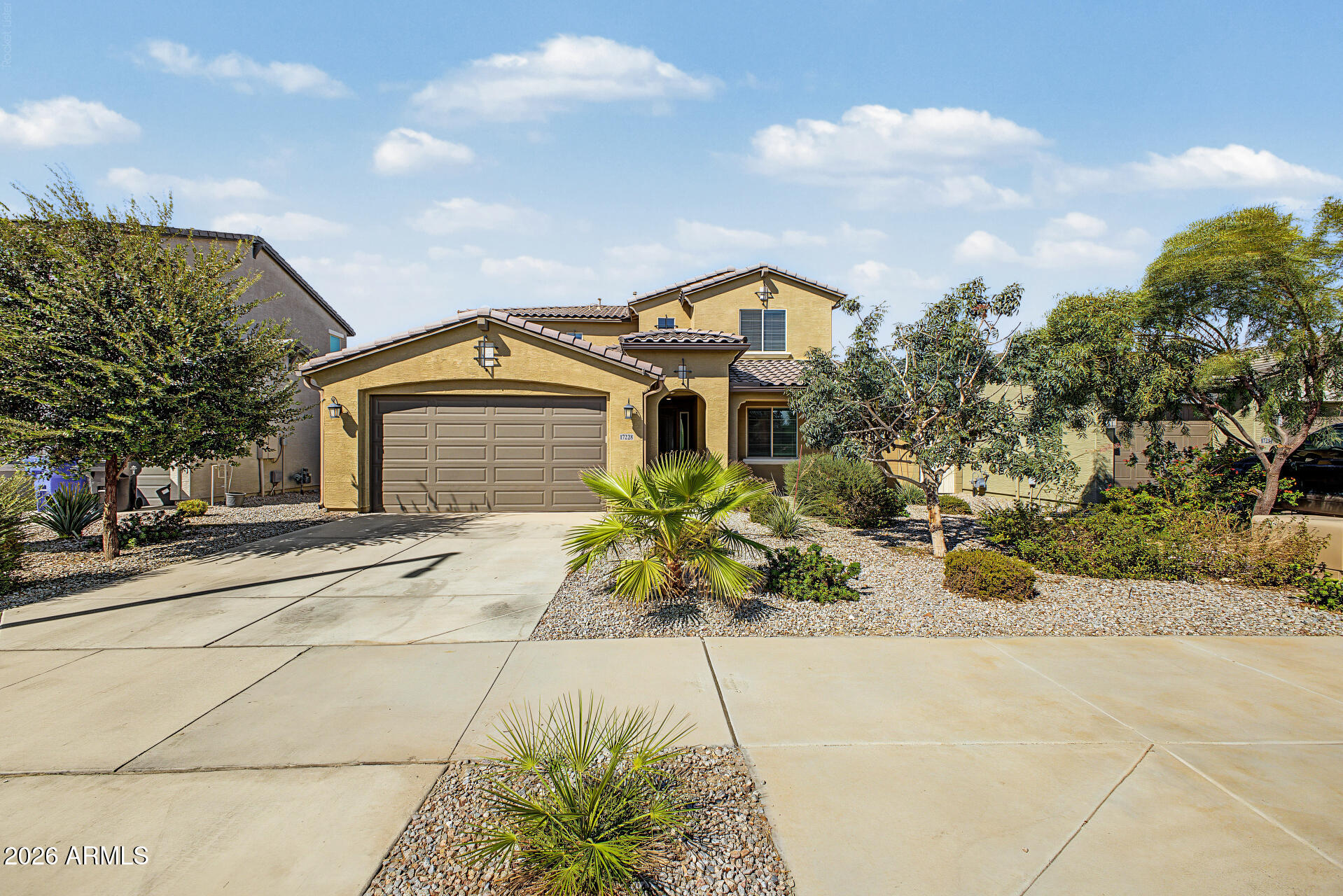 Two-story tan stucco home with a double garage, wide driveway, and desert landscaping.