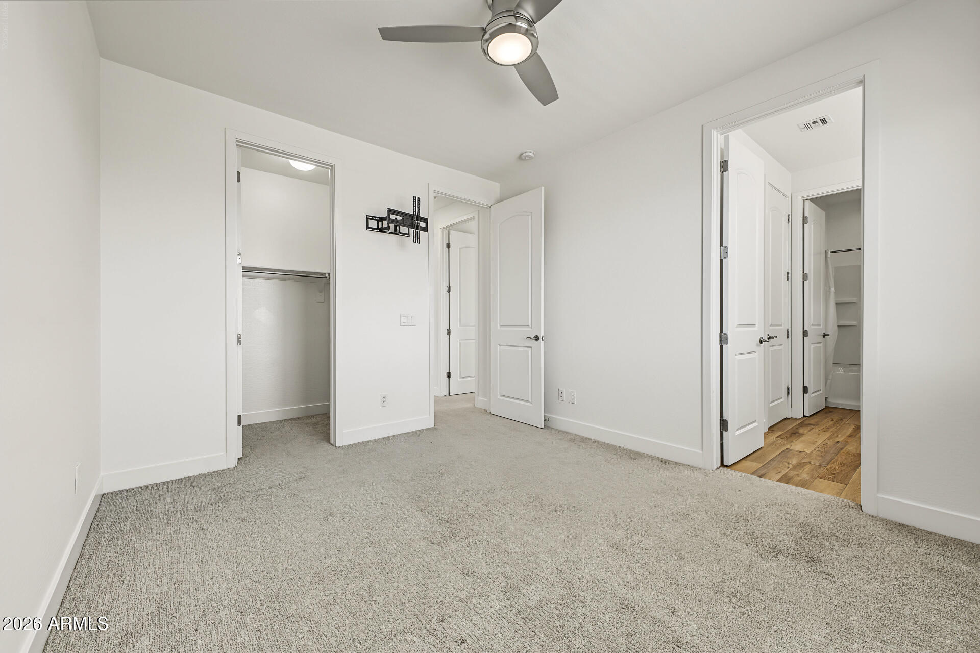 Empty carpeted bedroom with a ceiling fan and open doorways to closets and an adjoining wood-floored area.