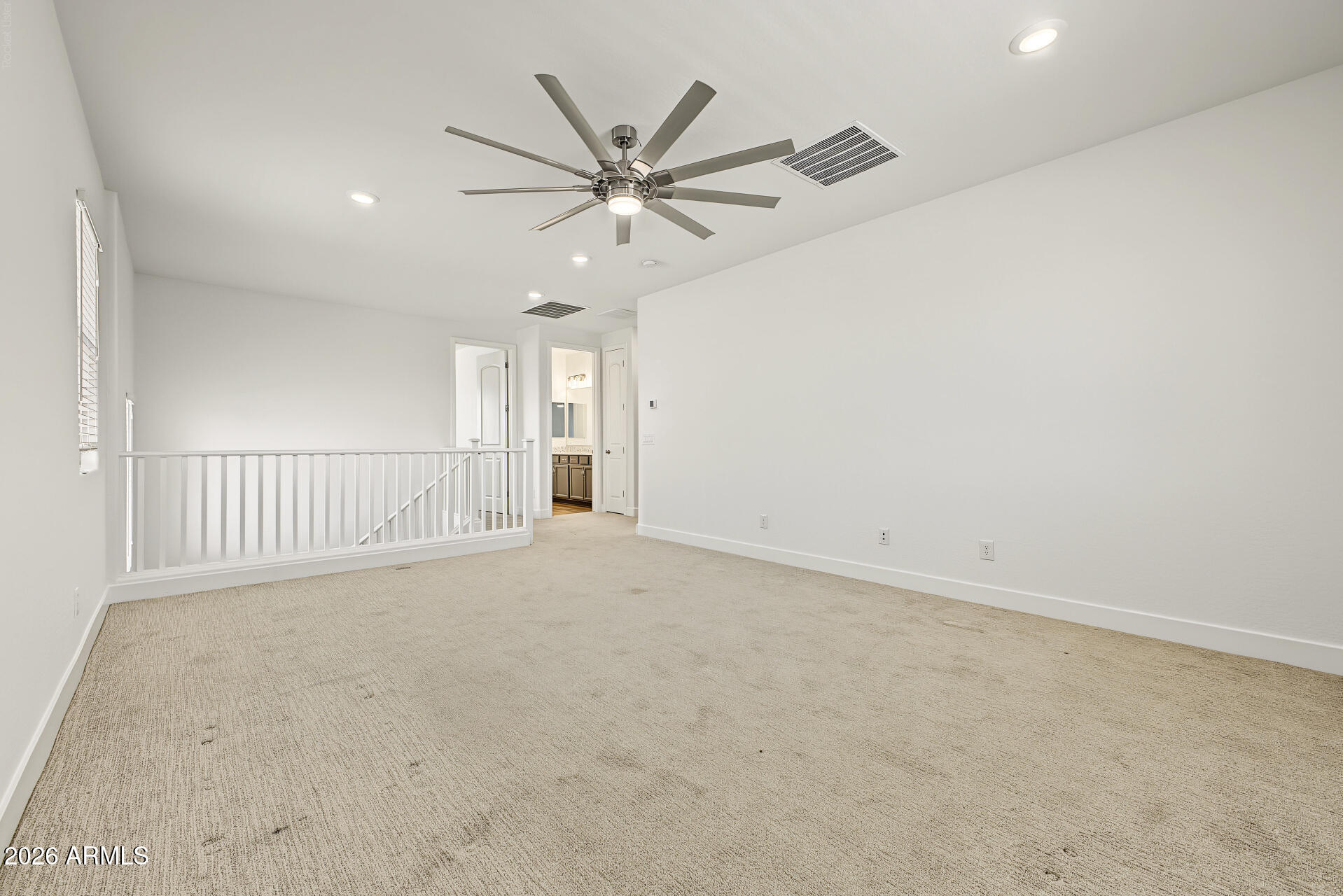 Empty carpeted upstairs room with a large ceiling fan and railing overlooking the stairs.