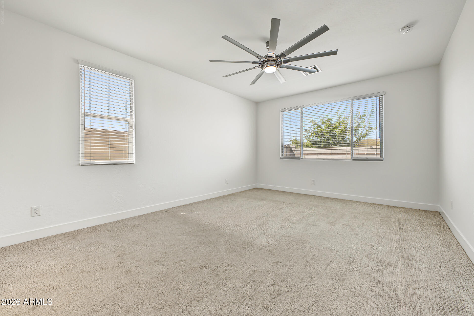 Empty bedroom with beige carpet, two windows with blinds, and a modern ceiling fan.