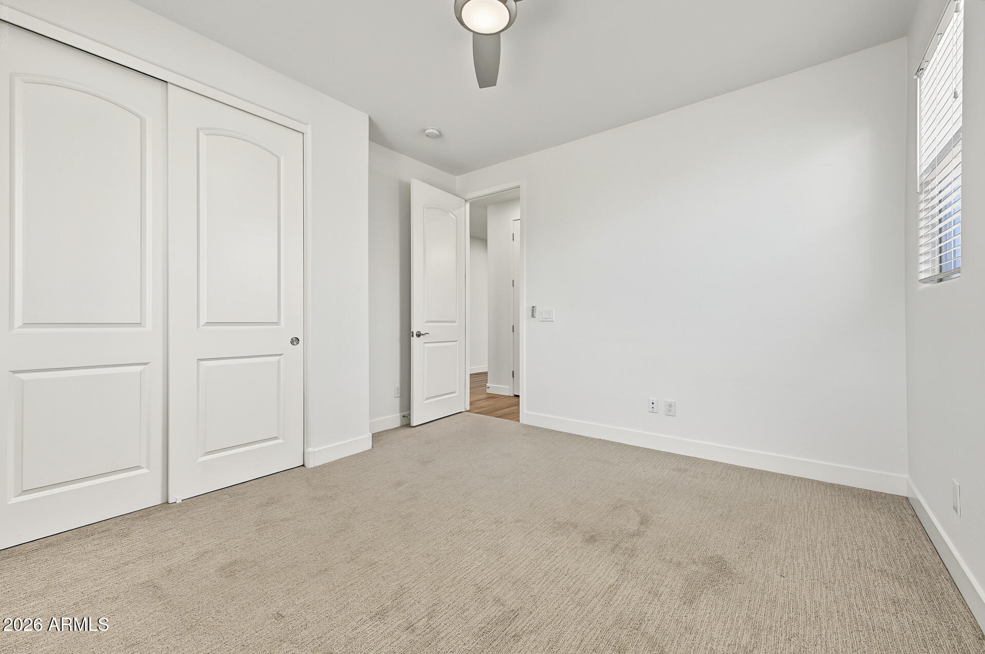 Empty bedroom with beige carpet, paneled closet doors, a window with blinds, and a ceiling fan.