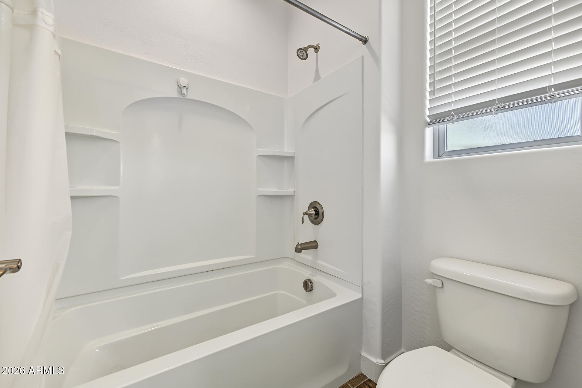 White bathtub and shower with molded shelves beside a toilet and window with blinds.