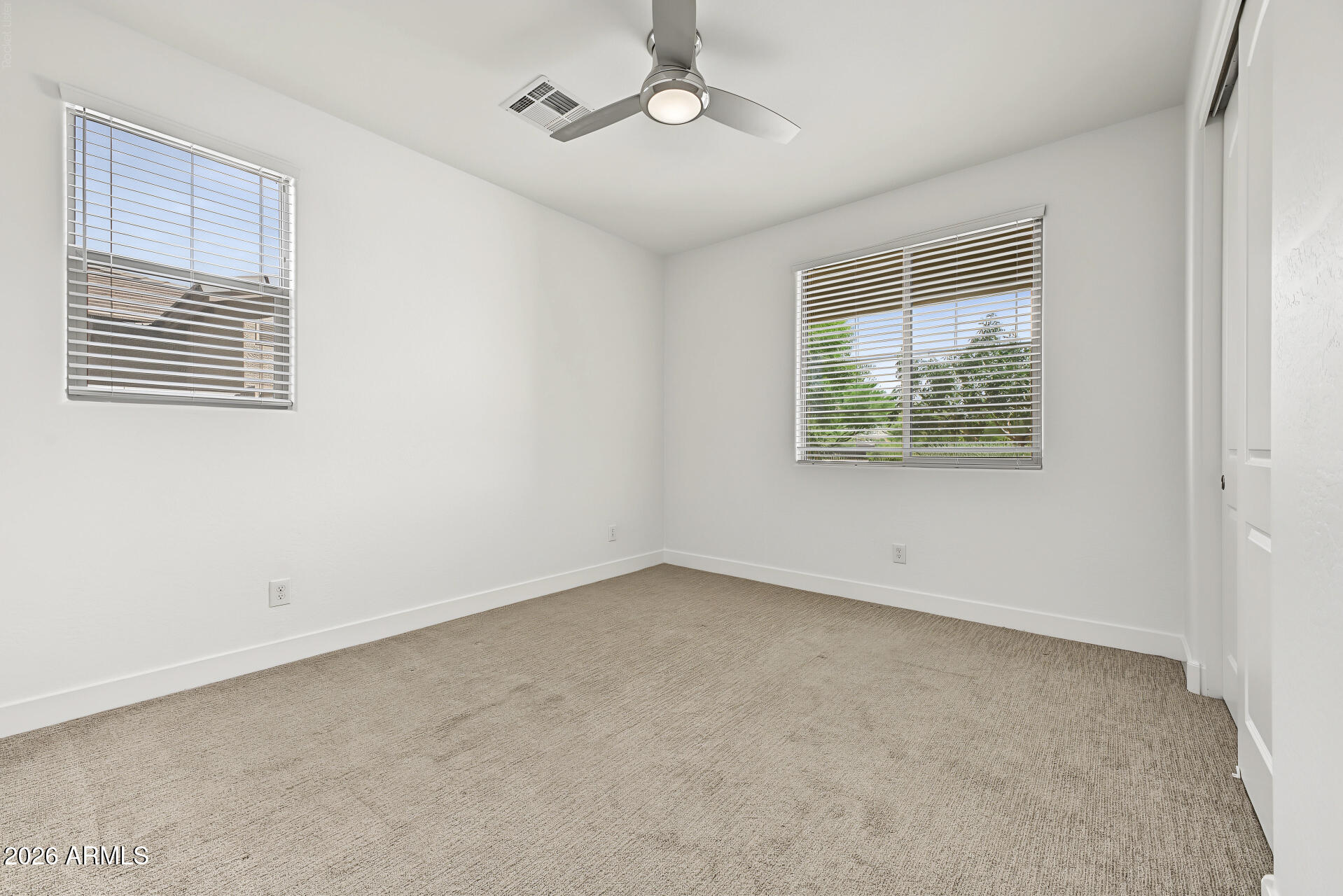 Empty bedroom with beige carpet, two windows with blinds, and a ceiling fan.