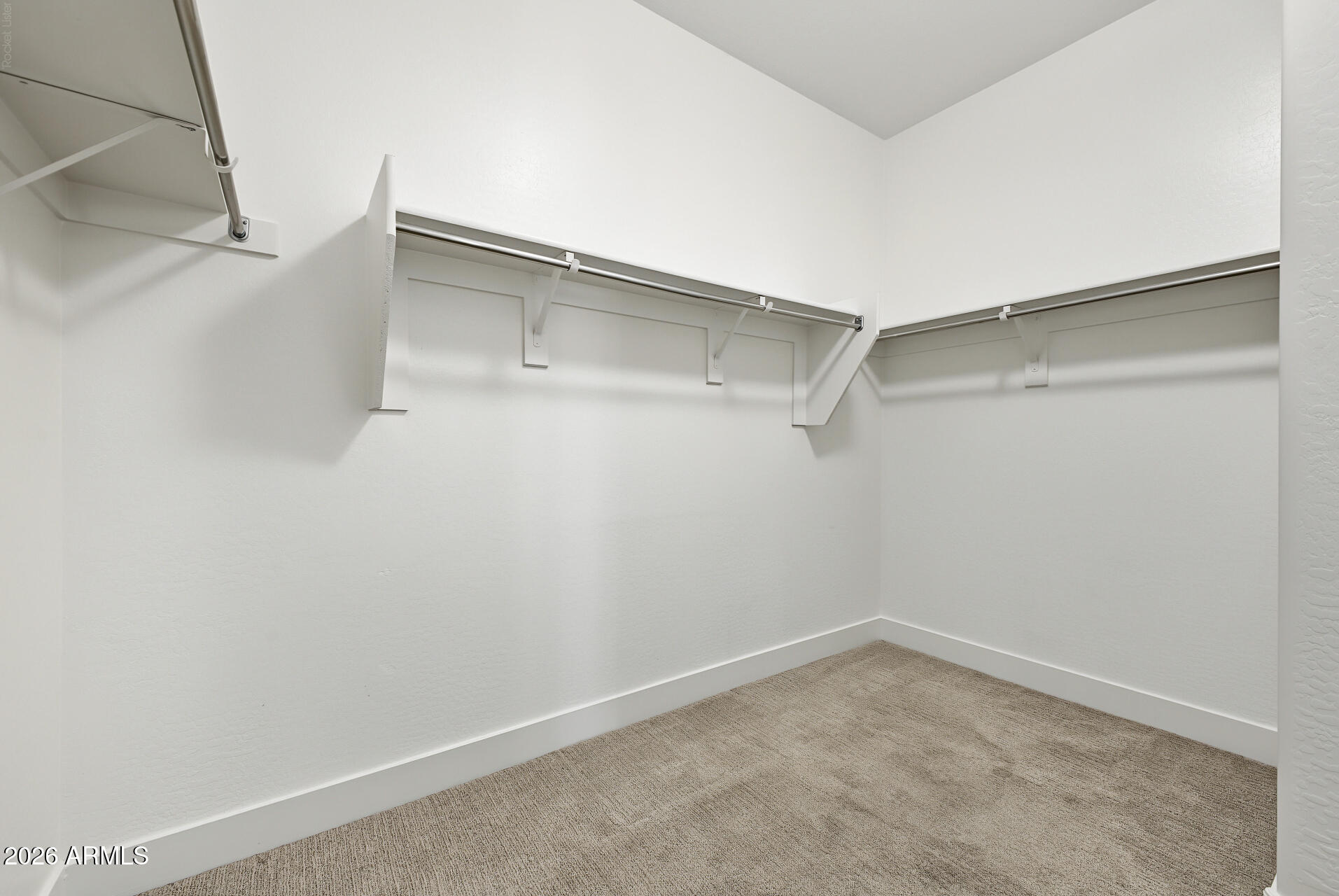Empty walk-in closet with beige carpet and wraparound shelves and hanging rods.