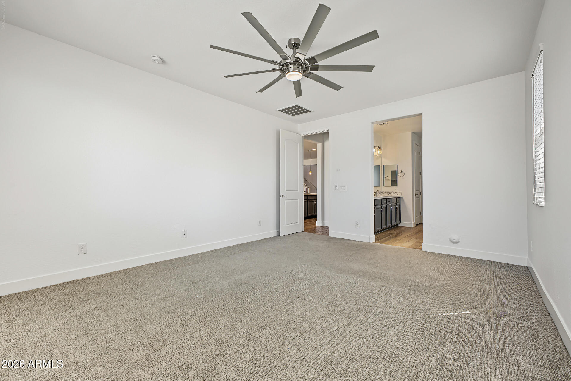 Empty bedroom with beige carpet, a large ceiling fan, and doorways to the bathroom and hall.