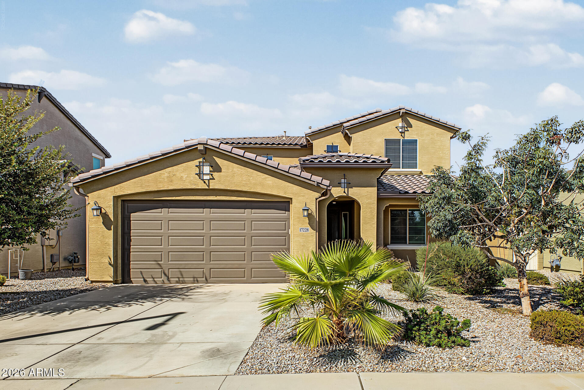 Two-story tan stucco home with a two-car garage, arched entry, and desert landscaping.