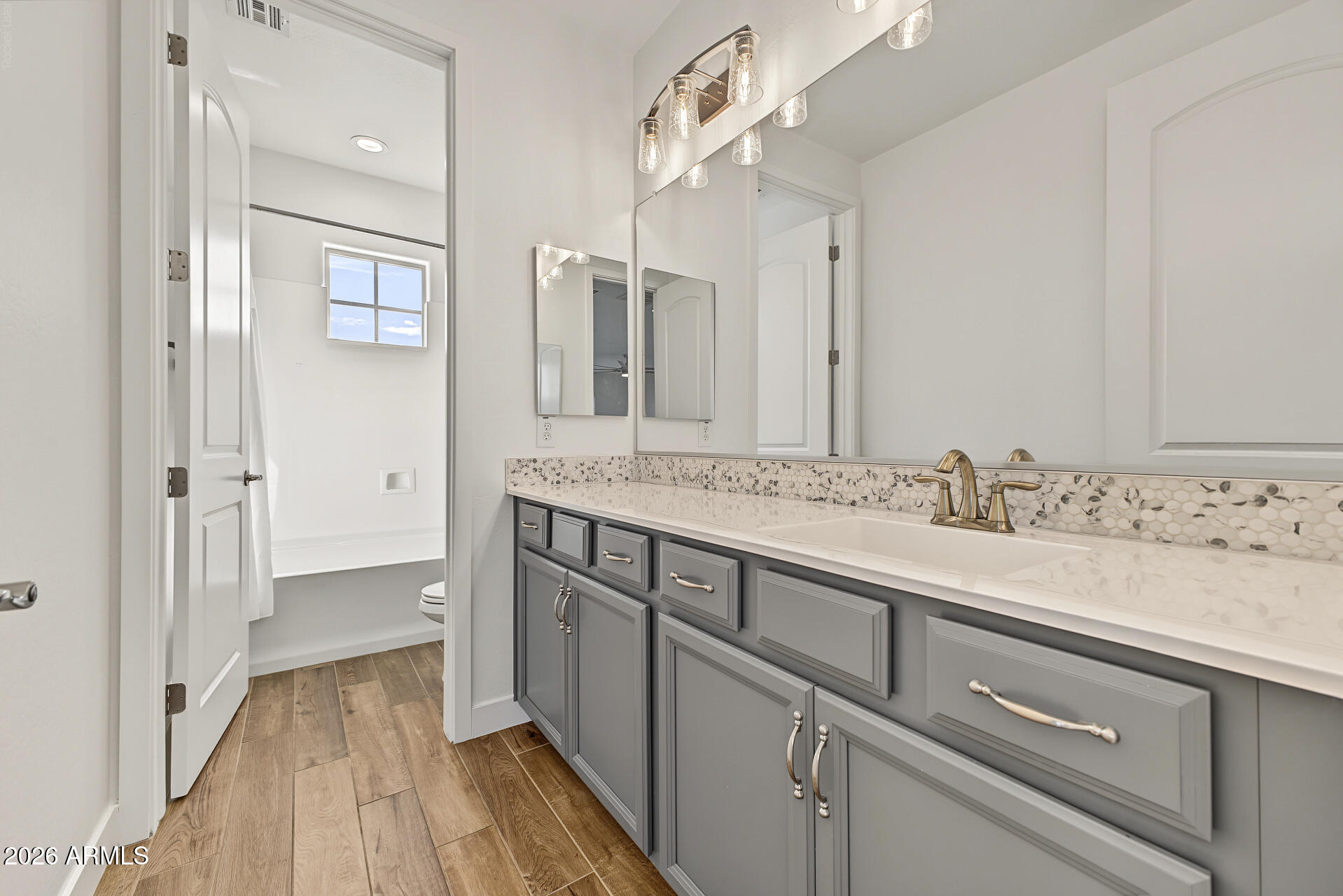 Bathroom with a long gray vanity, mosaic backsplash, brass fixtures, and a tub beyond the doorway.