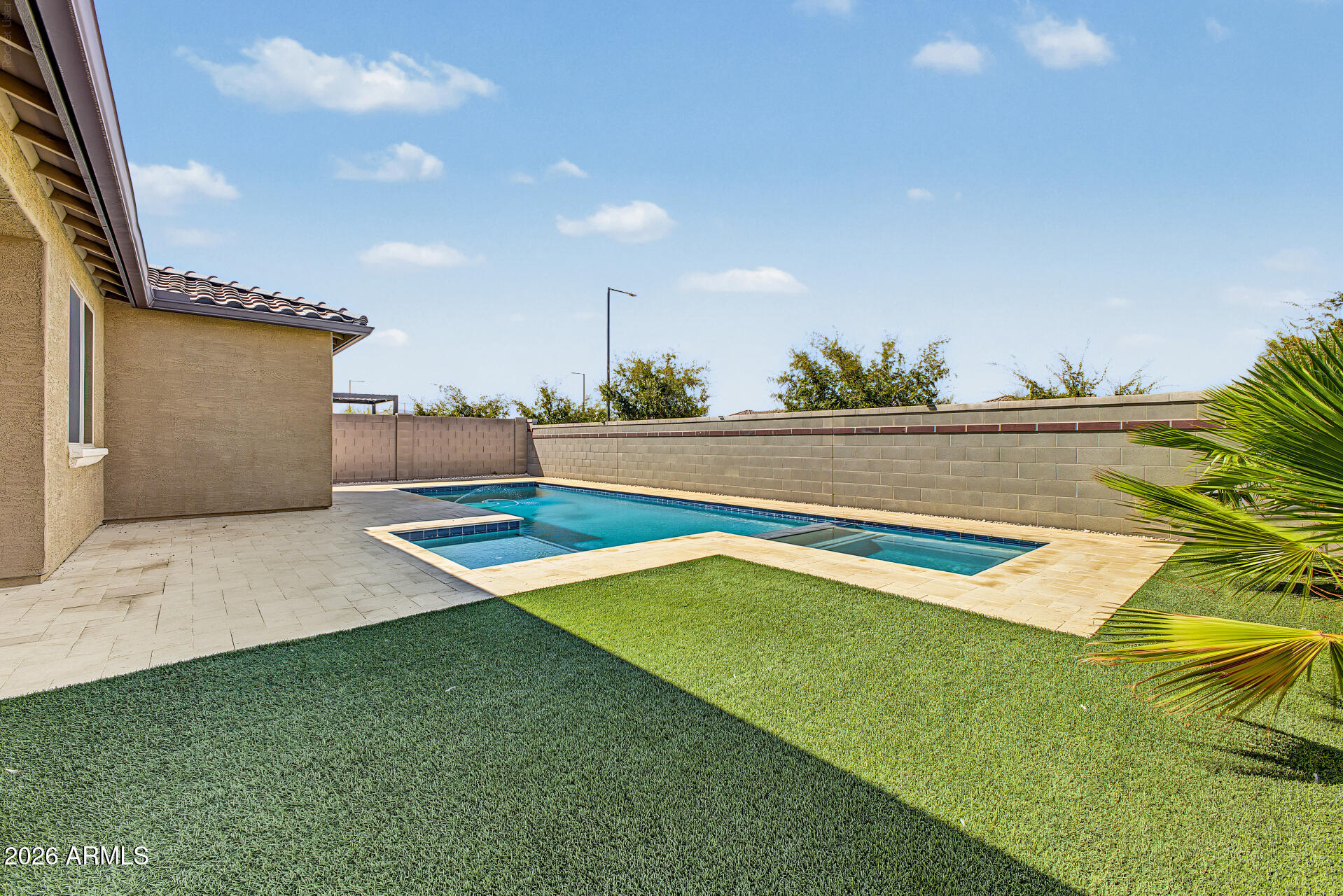 Backyard pool beside a tan stucco home, with a stone patio and green lawn.