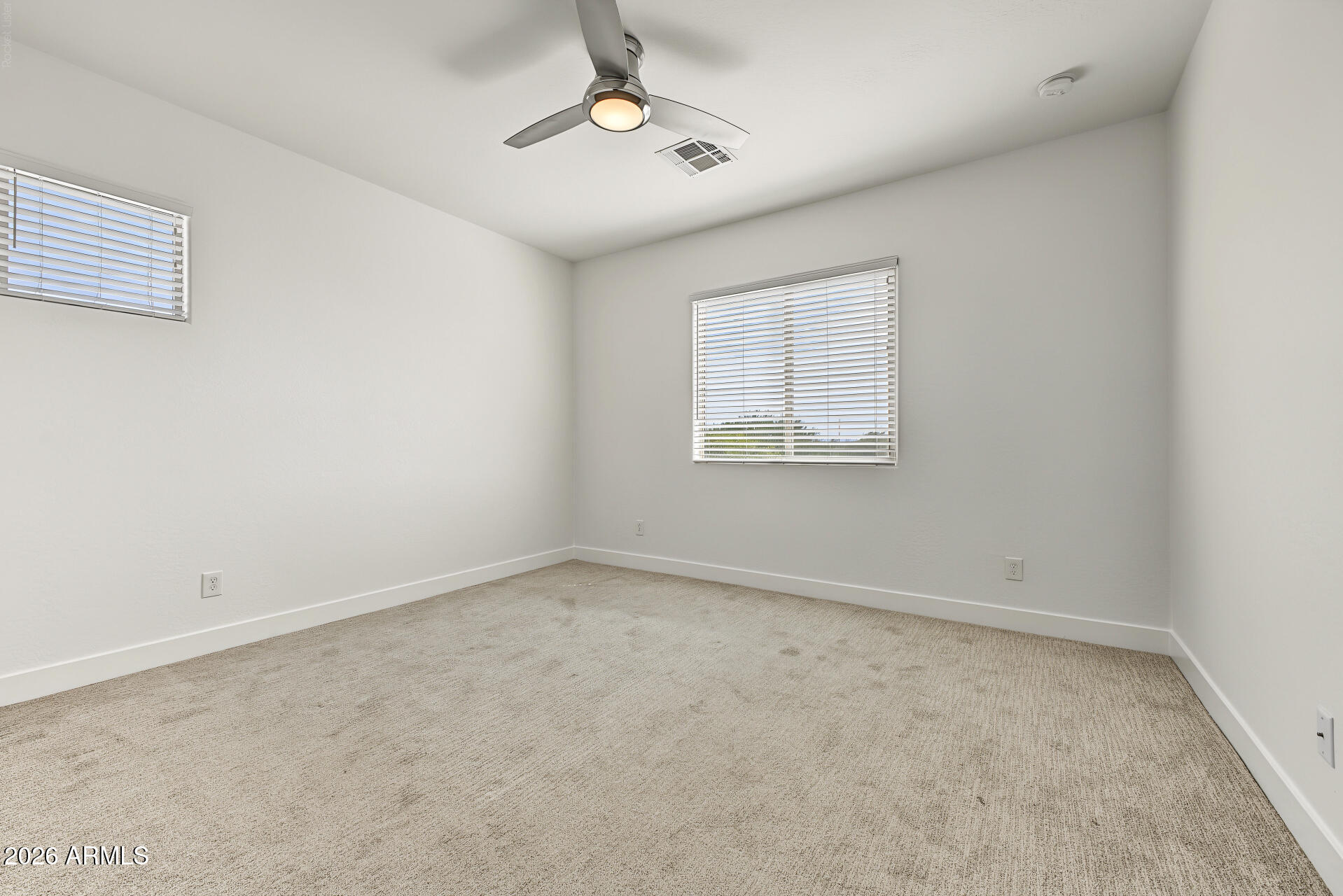Empty bedroom with beige carpet, two blind-covered windows, and a ceiling fan.