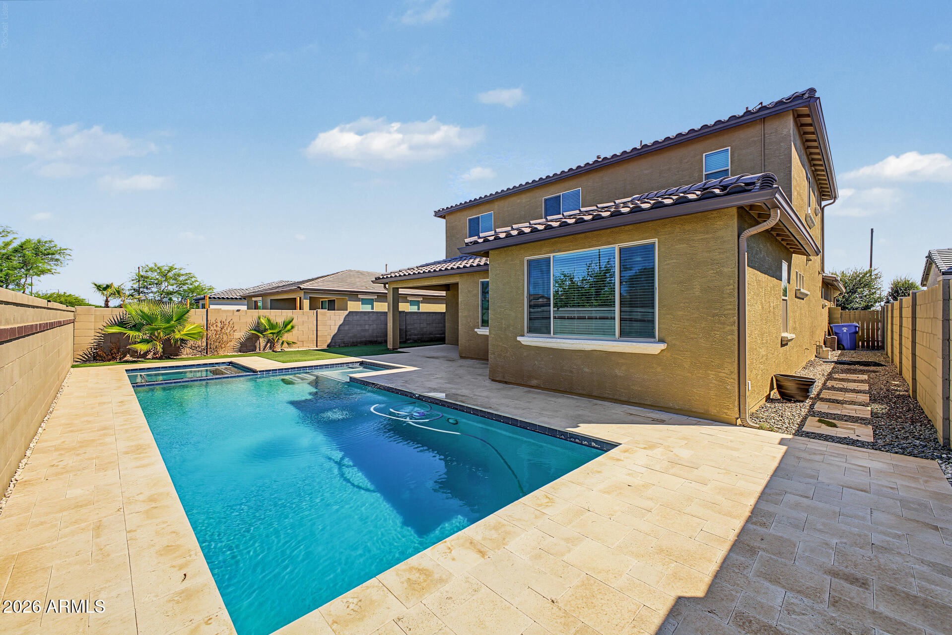 Backyard pool beside a two-story tan stucco home, with a broad paved deck and block walls.