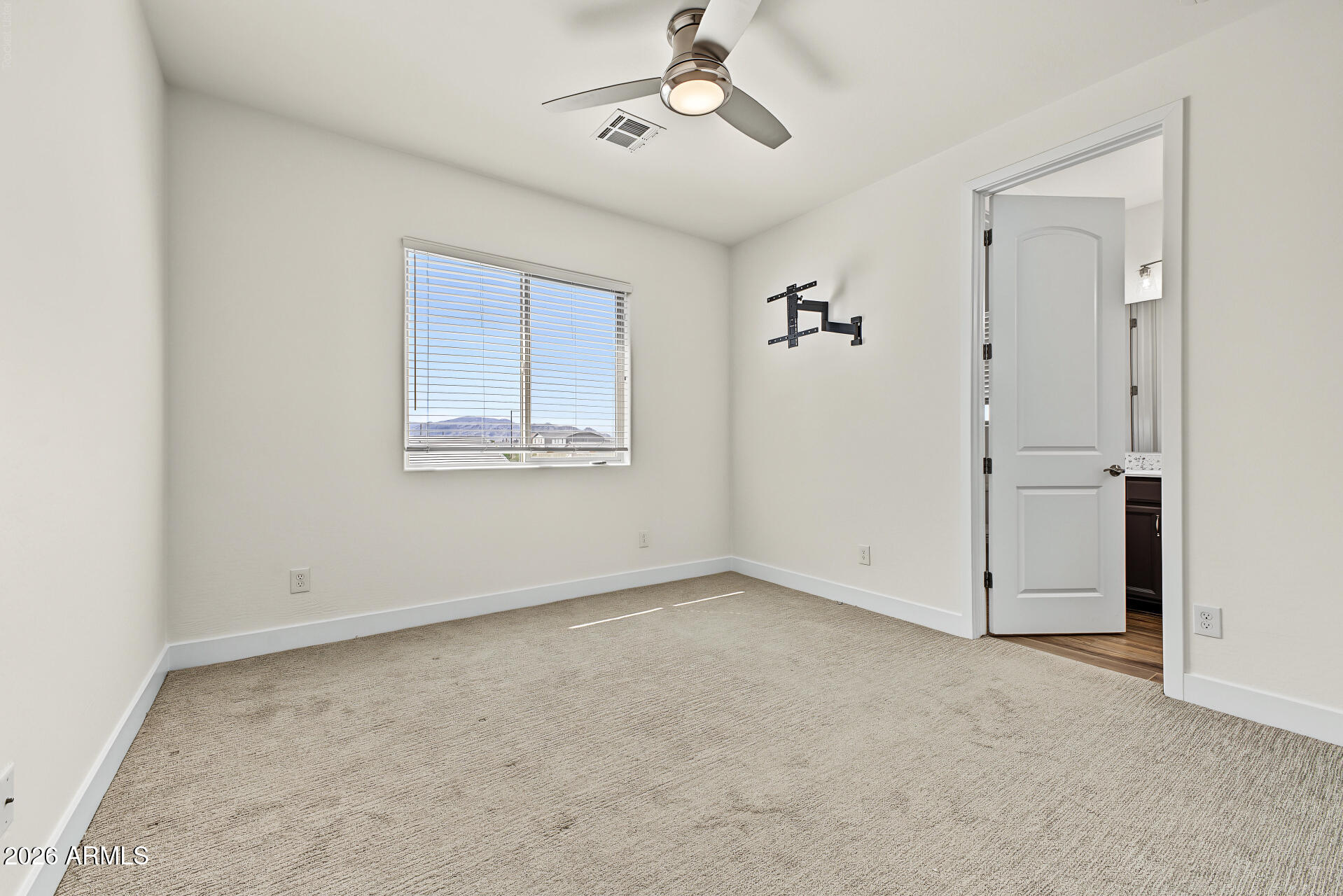 Empty carpeted bedroom with a ceiling fan, window blinds, and a wall-mounted TV bracket.
