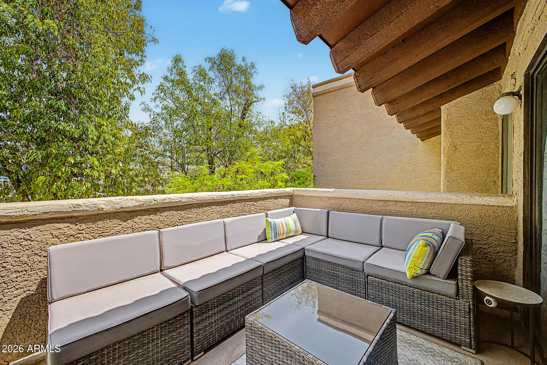 Furnished balcony with a gray sectional and glass-top coffee table overlooking leafy trees.