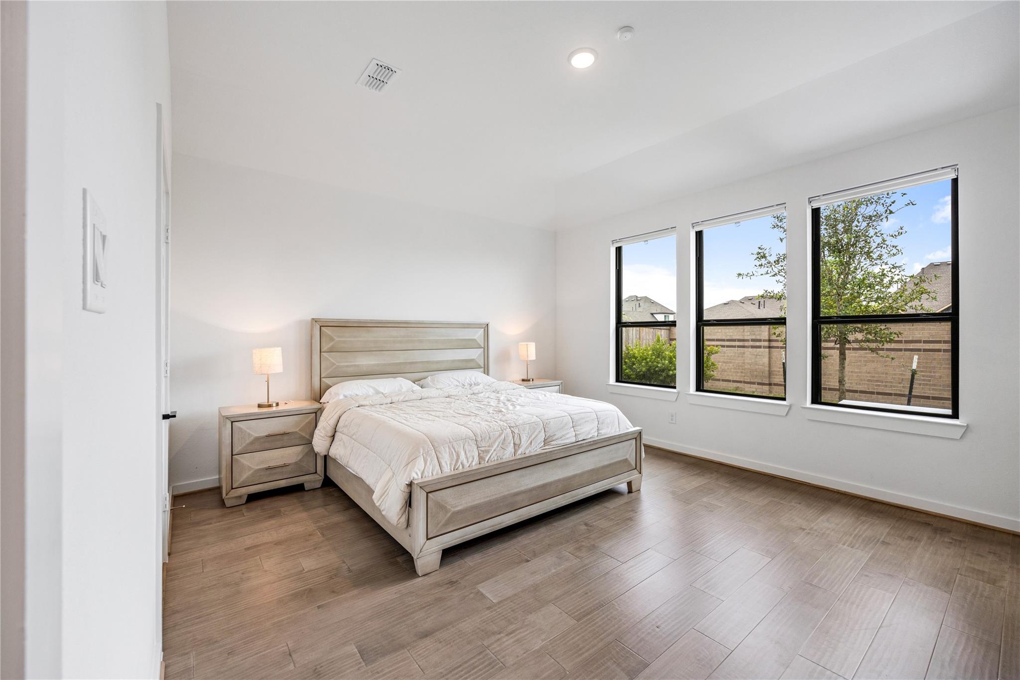 Staged bedroom with a light wood bed, white bedding, and three tall windows overlooking a fenced yard.