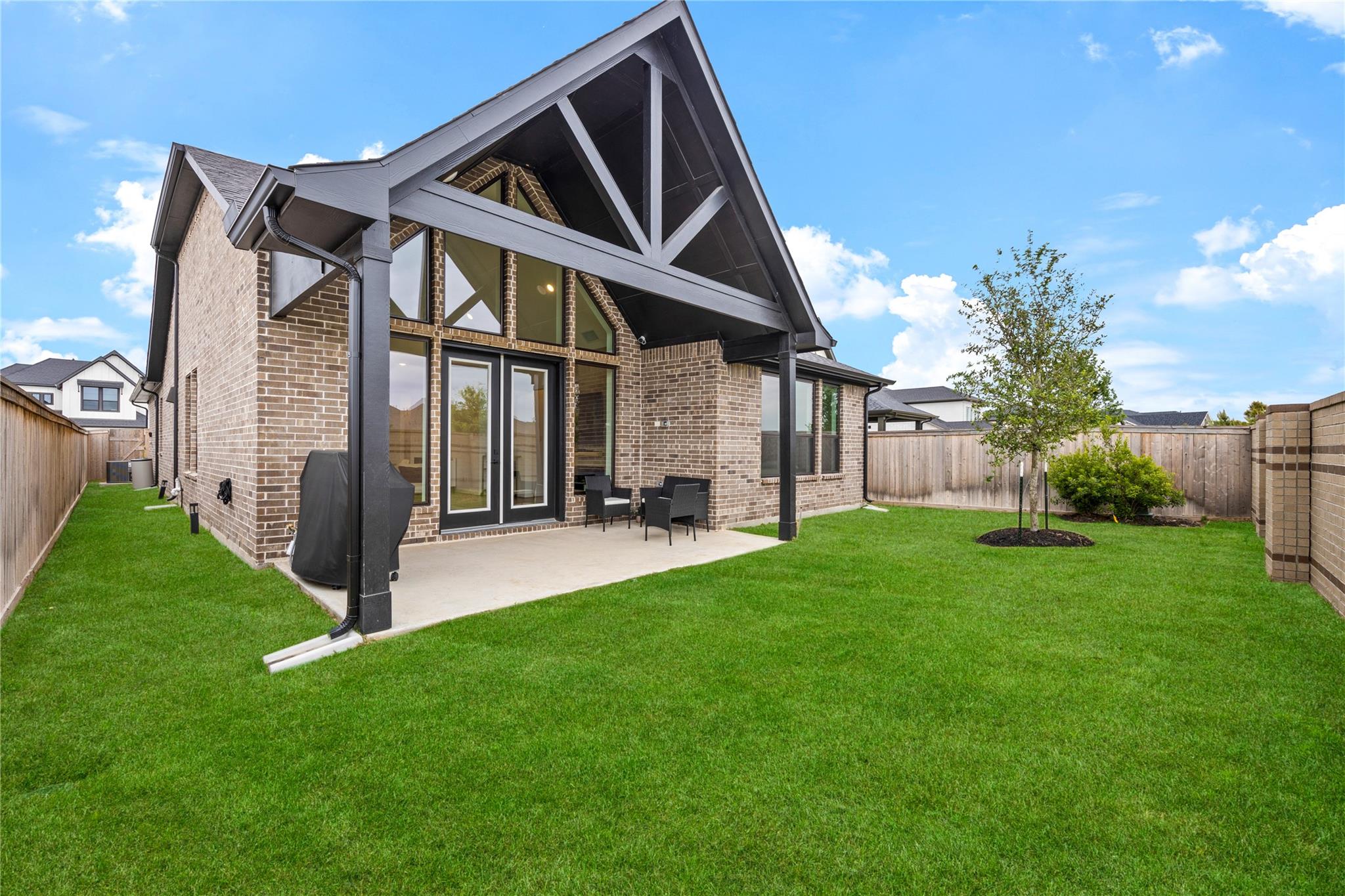 Brick home’s rear patio beneath a tall gable, overlooking a fenced backyard with a broad lawn and young tree.